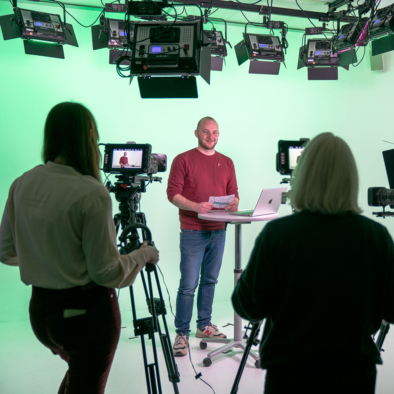 Person steht mit Moderationskarte im Filmstudio, zwei Personen davor stehen an Kameras.